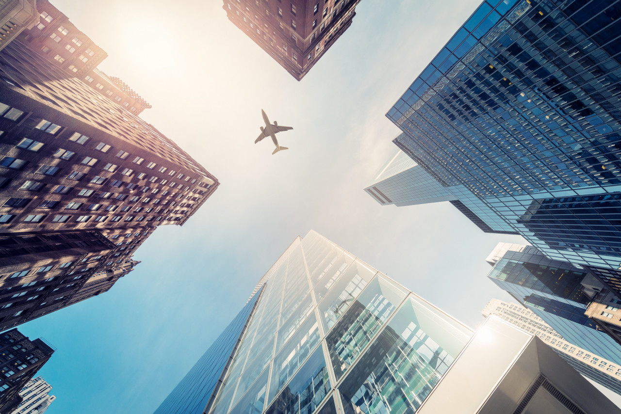 A view of an airplane flying in the sky between buildings.