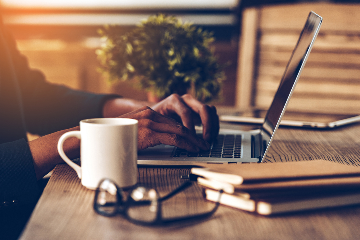 Fingers typing on a laptop keyboard atop a desk with a coffee cup, glasses and notebooks near by.