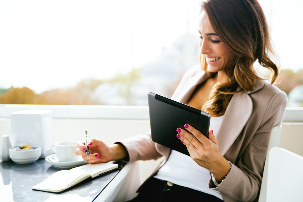 A woman sitting outside at a restaurant while holding her Ipad and taking notes over her financials.