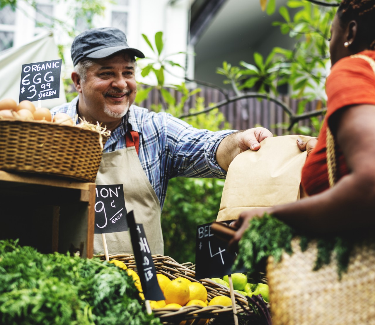 A man handing a customer her purchase at a farmer's market on a sunny day.
