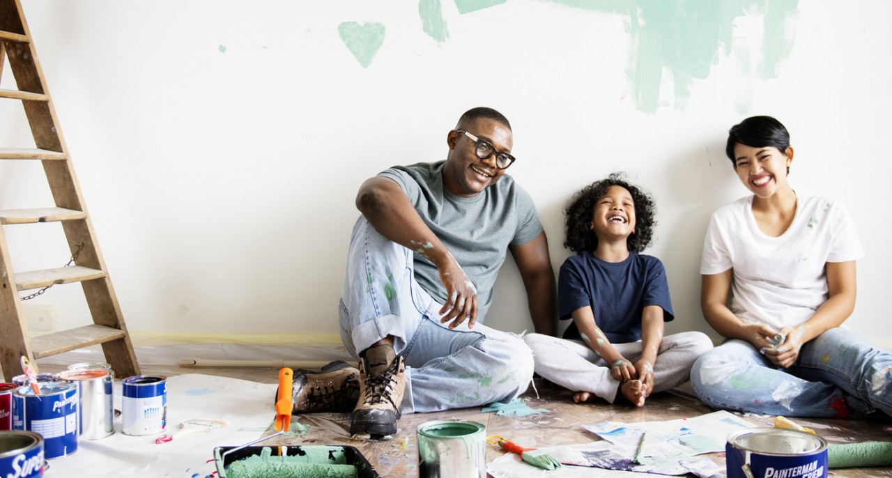 A family smiling while sitting on the floor of an empty, unfinished room with paint cans, brushes, tarps and a ladder.