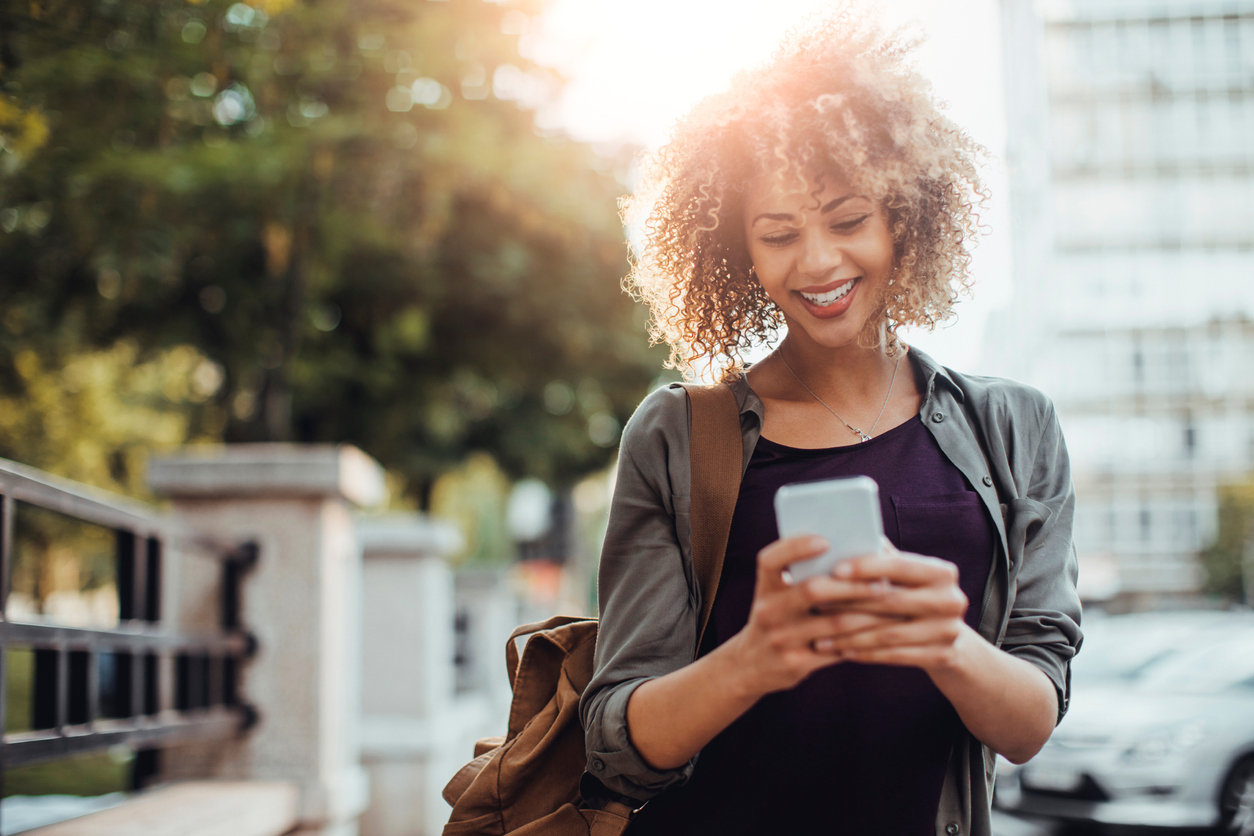 A woman in the city looking at her phone while smiling on a sunny day.