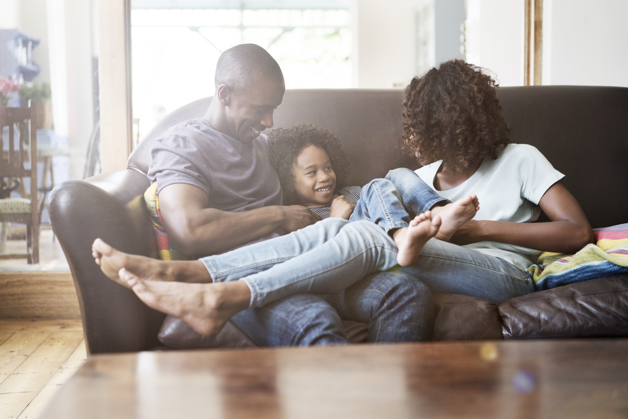 A family at home on the couch with their legs over the tops of one anothers smiling while enjoying quality time together.