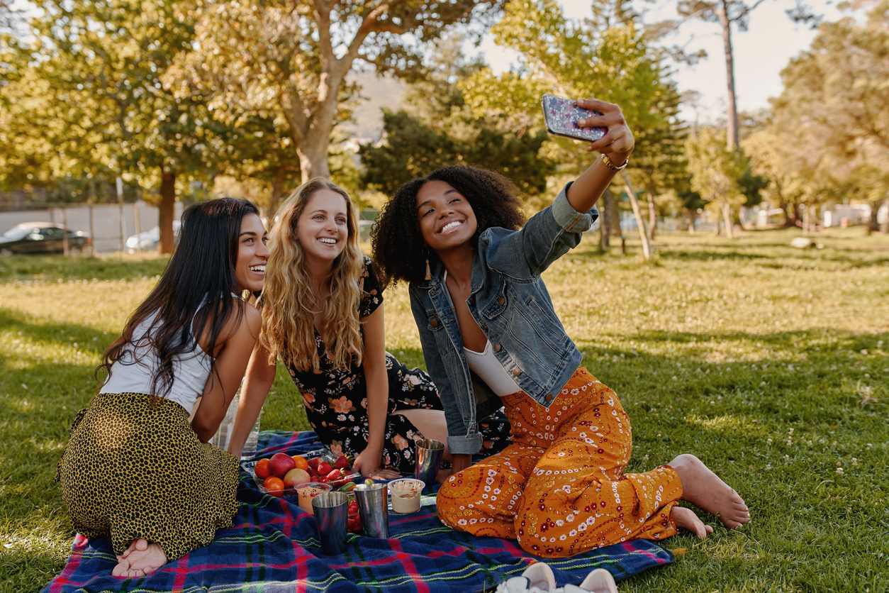 A group of happy females sitting together on a blanket in the grass enjoying a picnic in the park while taking a selfie.