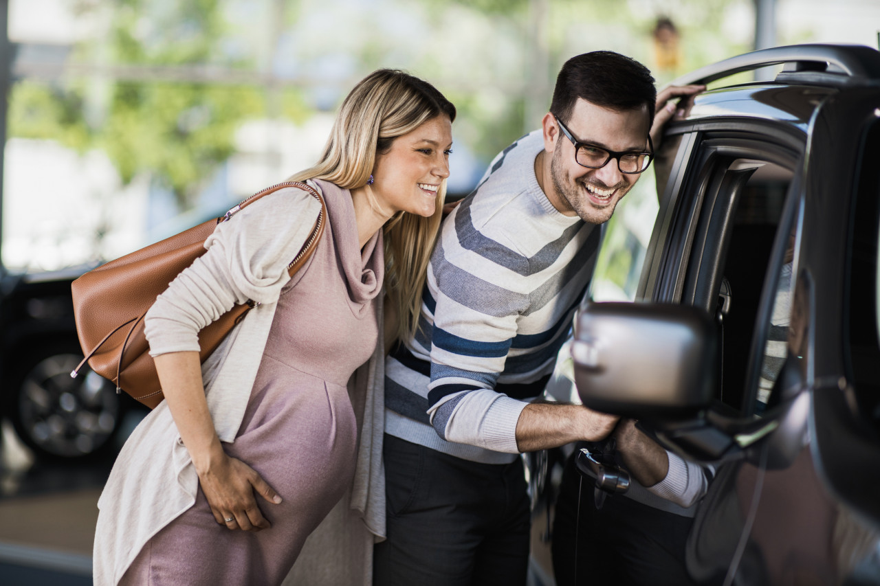 A pregnant couple looking into the window of a brand new car.