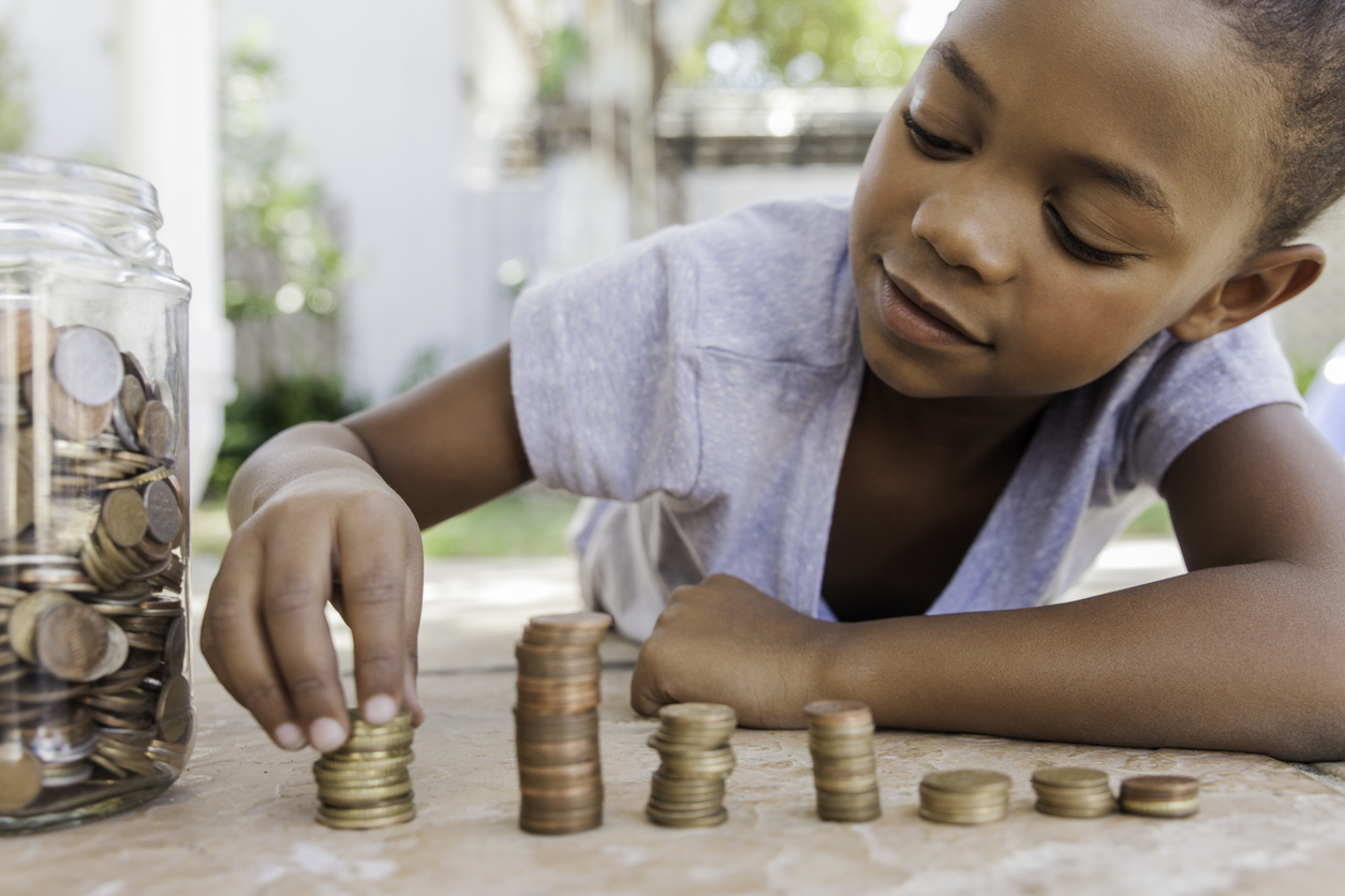 A child laying on her stomach with a full jar of coins next to her as she is counting while stacking piles of coins up on a hard surface.