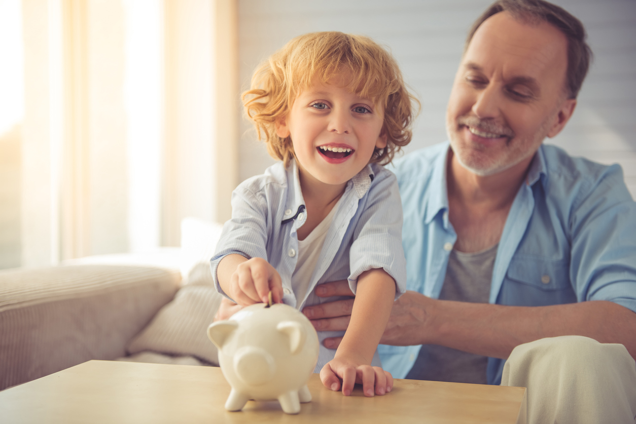 A child happily inserting a coin into a piggy bank as his father proudly watches.