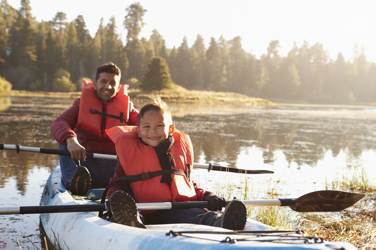 A father and son in a kayak wearing life jackets on the water smiling with paddles in their hand.
