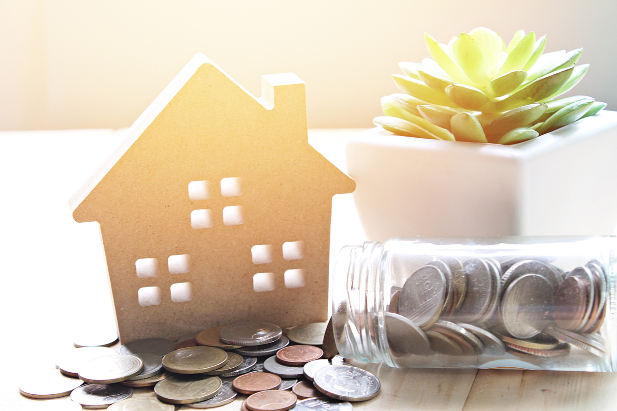 A cardboard cut out of a home sitting on a table with a jar of money tipped over and the coins pouring out.