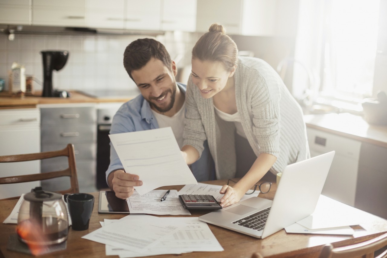 A couple at their dining table going over financials on their paper applications and laptop while using a calculator.