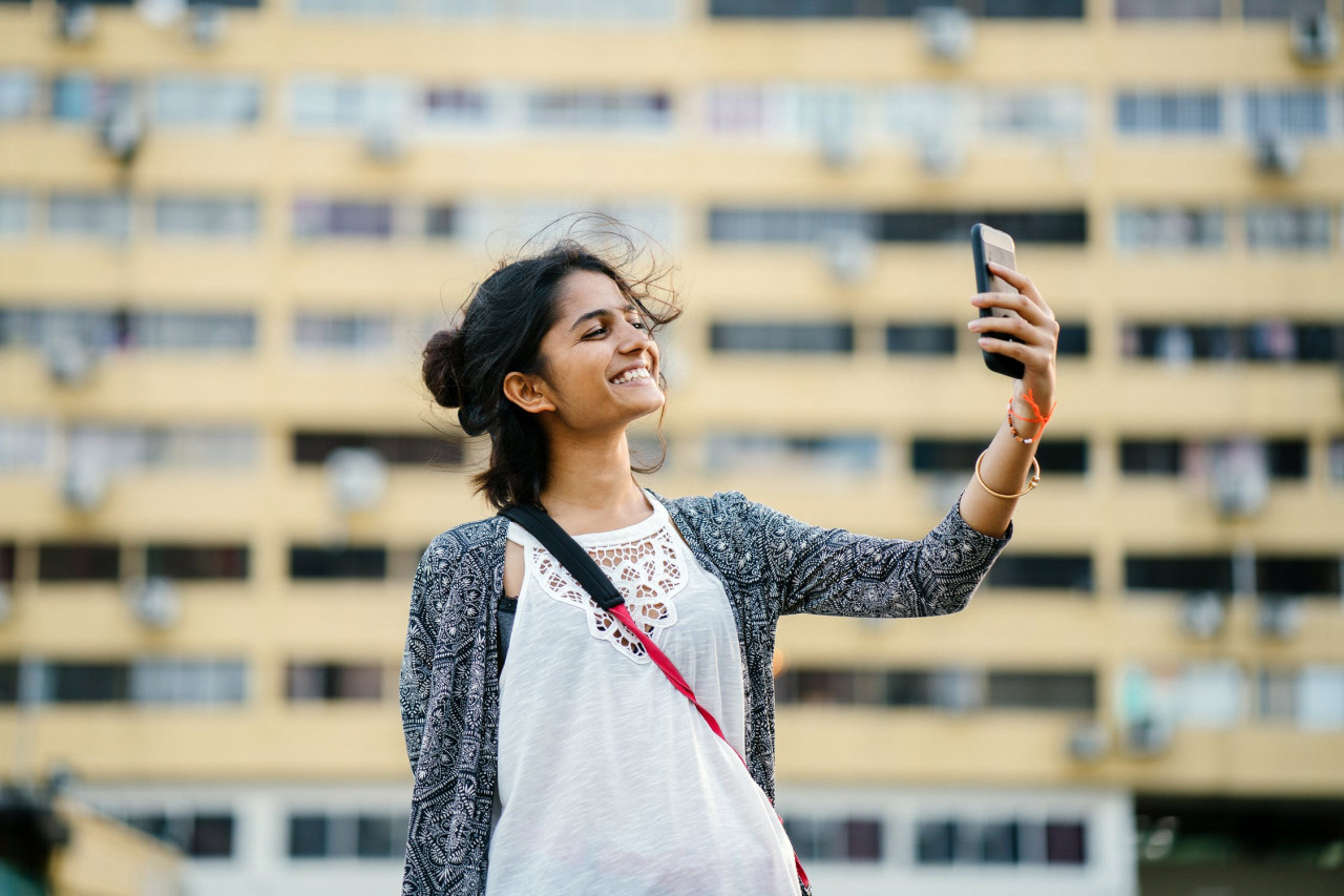 A woman outside in a city holding her phone up and smiling at it.
