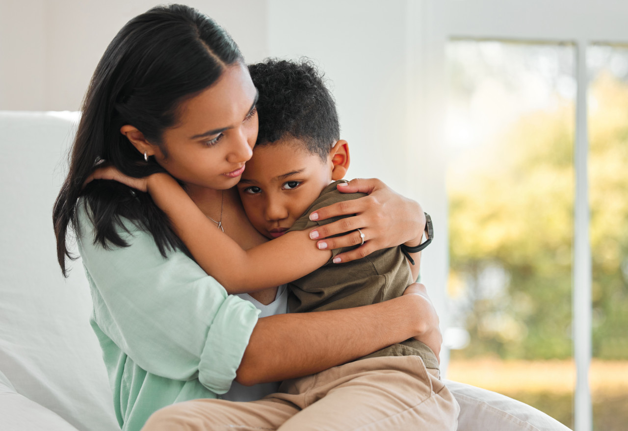 A young boy hugging a woman inside a home by a window.