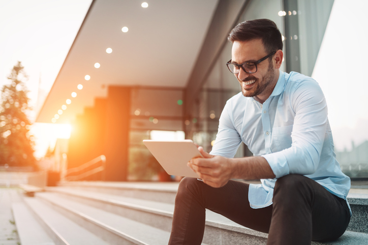 A man is sitting outside on a step, looking at his tablet.