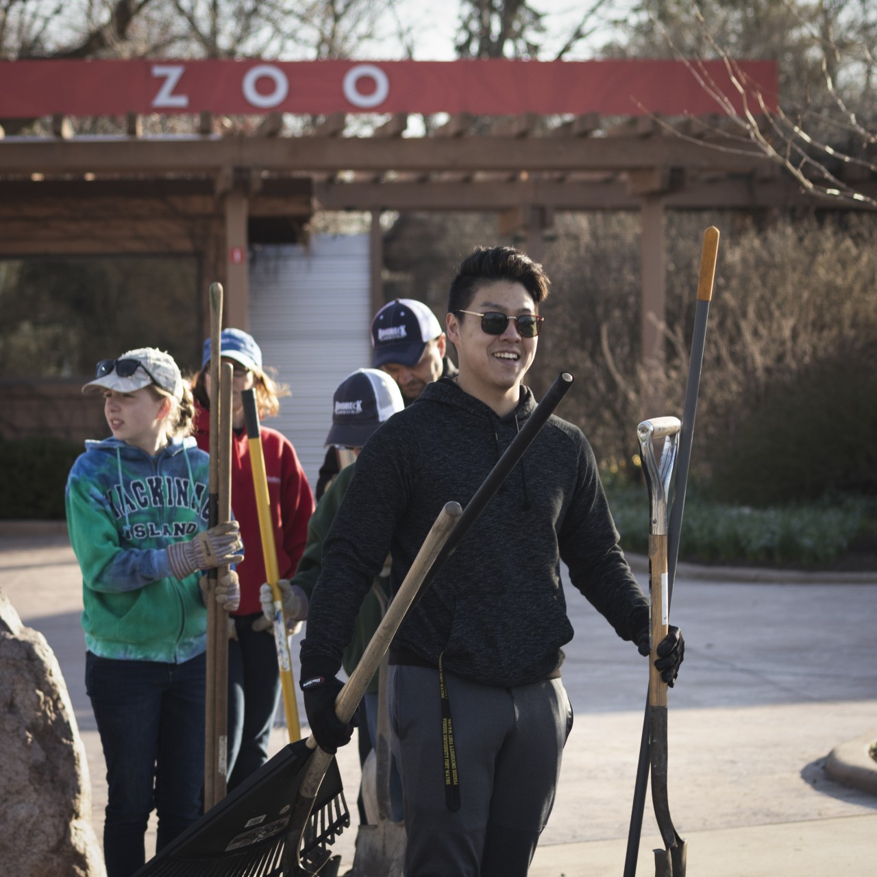 man smiling at camera while holding a number of landscaping tools
